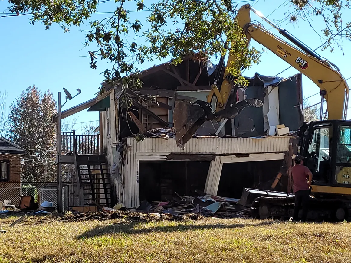 A house being demolished with an excavator in the foreground.